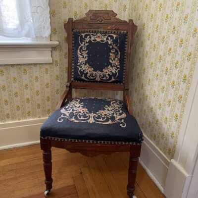 A modern color photograph of an artifact from the Parish House and Museum—a wooden chair with a padded seat and back, upholstered in embroidered fabric featuring a floral design on a navy blue background with white, red, and green flowers.