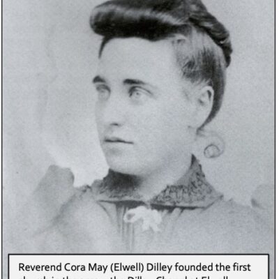 An old black and white close up photograph of Reverend Cora May (Elwell) Dilley. She is wearing a dark colored top with a small ribbon bow. Her hair is fashion in an updo with a large curl along her front hairline.