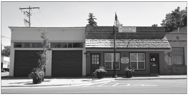 A black and white photo of a building with an attached two car on the left and a business on the right with two entrances.