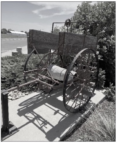 A black and white photo of a 1912 hose cart, featuring a metal frame with large wheels and a coiled fire hose, representing early 20th-century firefighting equipment.