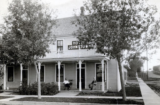 An old black and white photo of the two story Johnstown Hotel which has a balcony across the front and a porch underneath with two adults sitting in chairs.
