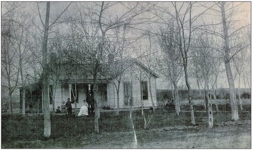 An old black-and-white photograph of the original Parish Family Homestead, a single-story house with a small front porch, surrounded by leafless trees, with the family standing in front.