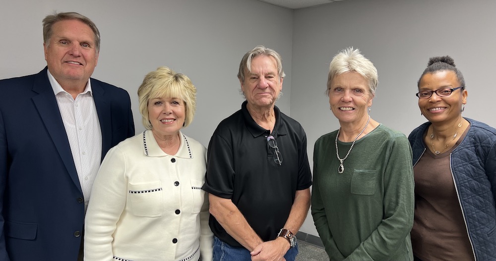 A group photo of five smiling people standing in a row against a gray wall.