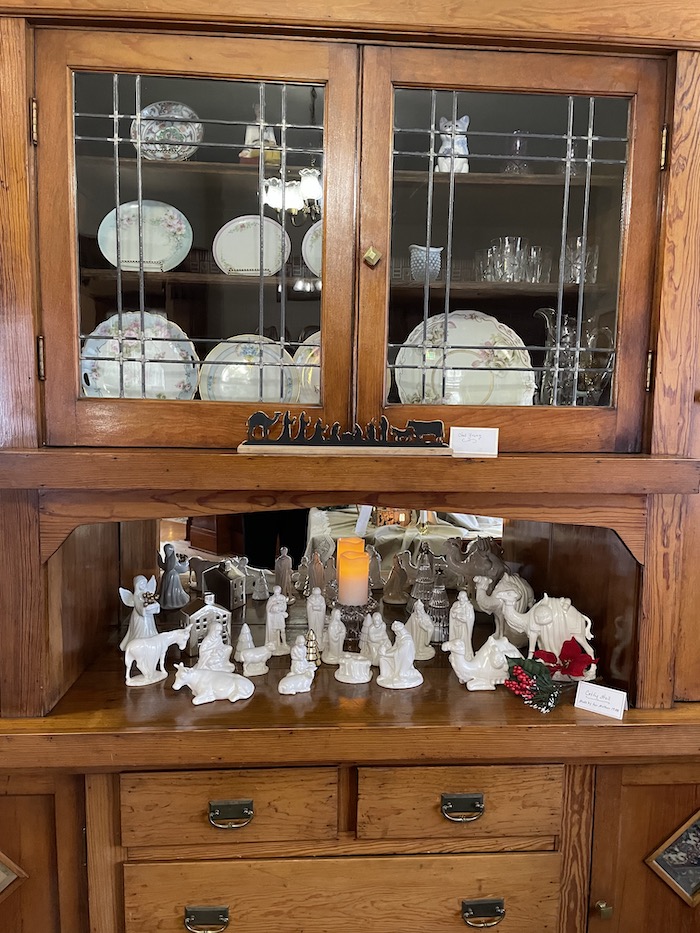 A built in wooden cabinet displays a large white nativity scene. A cabinet above has glass doors and displays antique china.