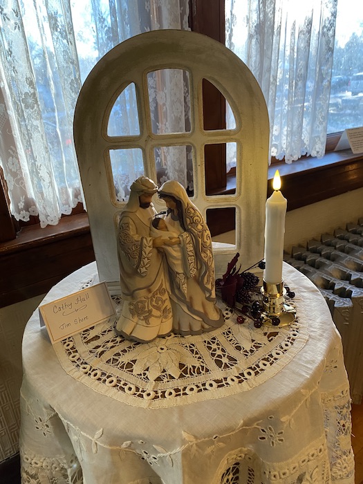 A nativity displayed on a small round table with a candle beside it and a decorative window standing tall behind it.