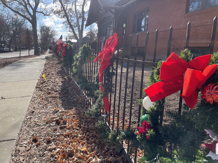 A black fence surrounding a brick home displays holiday garland and wreaths with red bows.
