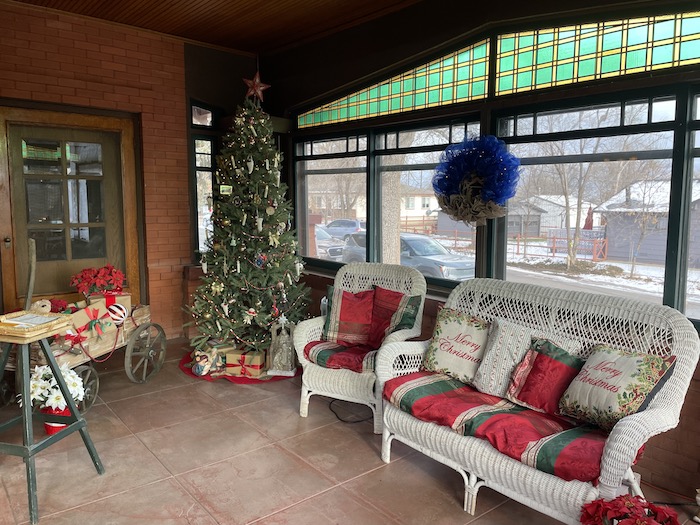 A large porch with green and yellow stained glass windows is decorated for the holidays. A white wicker patio set is adorned with red and green pillows with a decorated tree stands to its side.