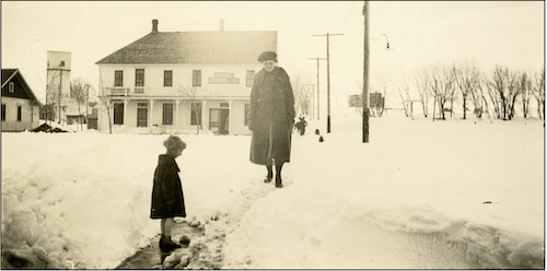 An old black and white photo of a woman and a child dressed in winter gear walking through deep snow.  An large white hotel building is in the background.