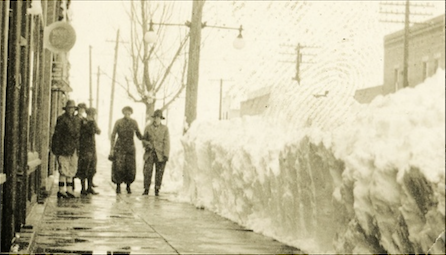 An old black and white photo of men and women walking down a sidewalk that has been shoveled. Along the entire street side of the sidewalk there is a large bank of snow nearly as tall as the men and women.