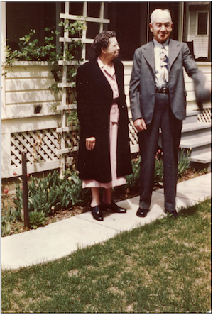 This photo shows an older man and woman standing on a sidewalk in front of the porch of a home.  She is wearing a dress and long coat and is looking to the side at her husband who is wearing a gray suit.