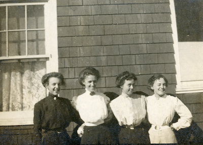 An old black and white photograph showing four women smiling and standing in a row with their arms around eachother.
