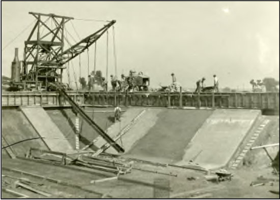 An older black and white photo showing a construction crew working on a pit with large construction equipment.