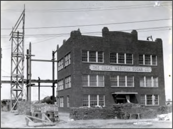 An older black and white photo of a 3 story brick building with 8 sets of windows and a large front entrance. A sign on the building reads, The Great Western Sugar Co.